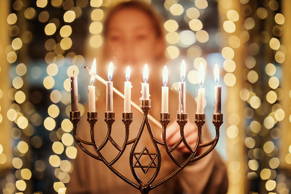 A hanukkiah in sharp focus being lit by a woman in the background.
